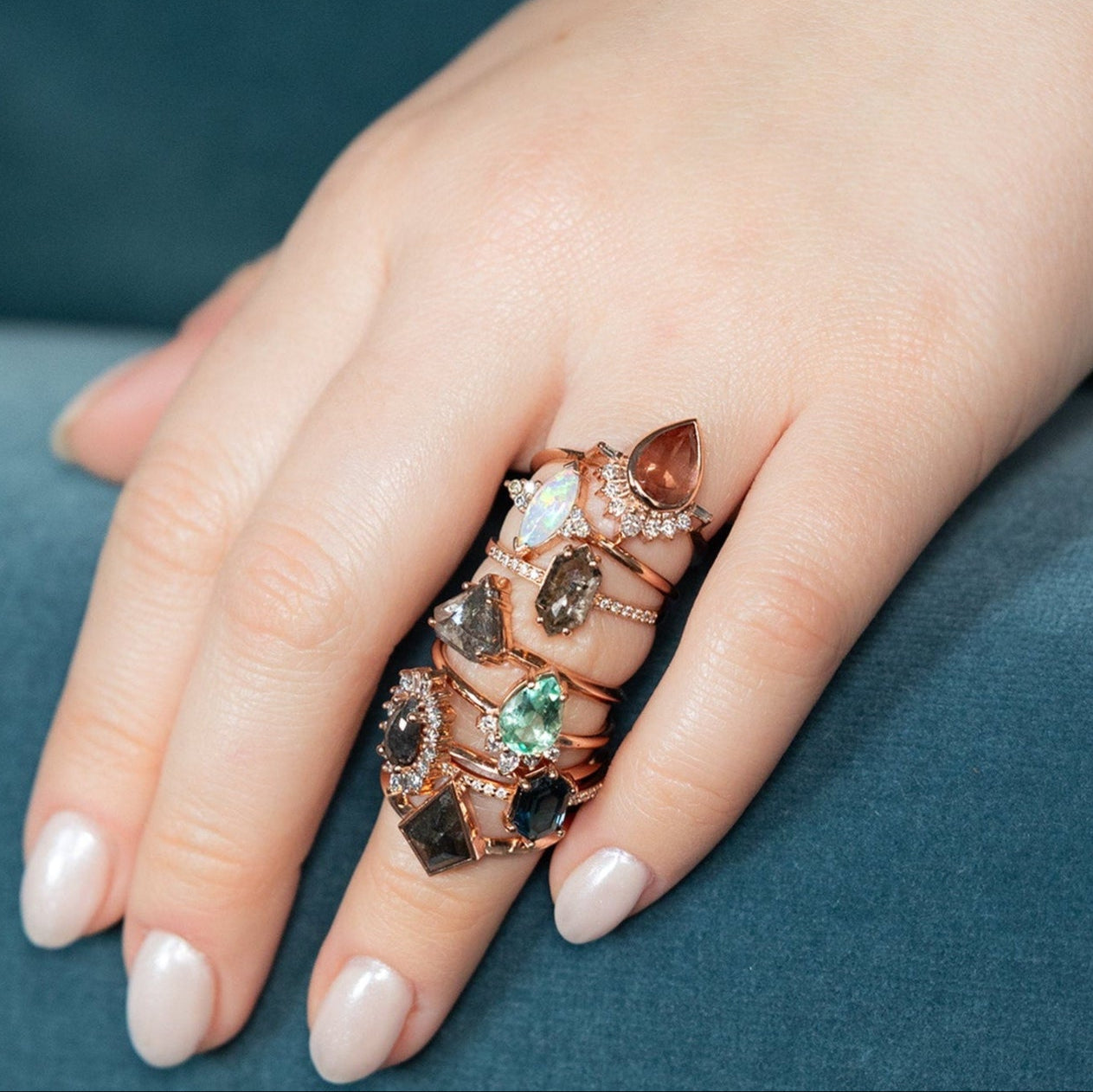 Model wearing seven diamond and gemstones engagement rings on ring finger with hand on blue velvet couch